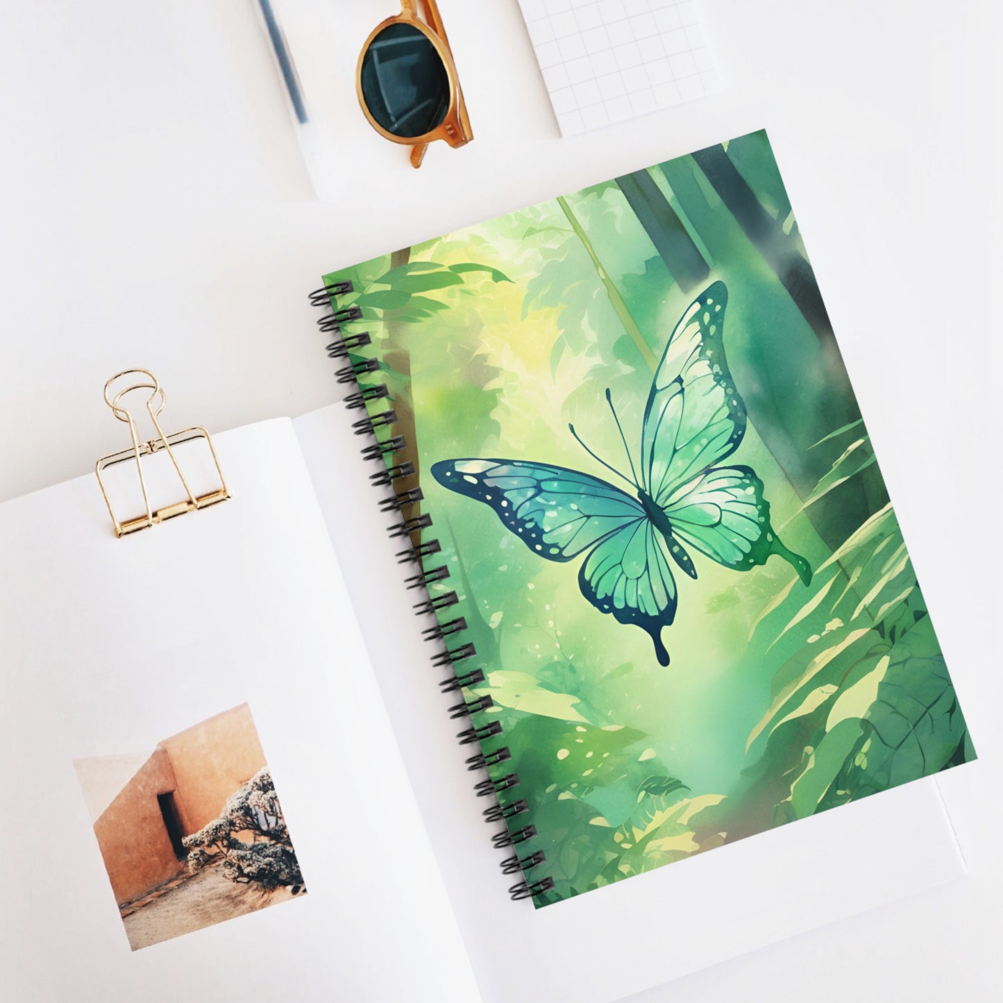 Notebook with a green butterfly design on a white surface with sunglasses and a photo clip.