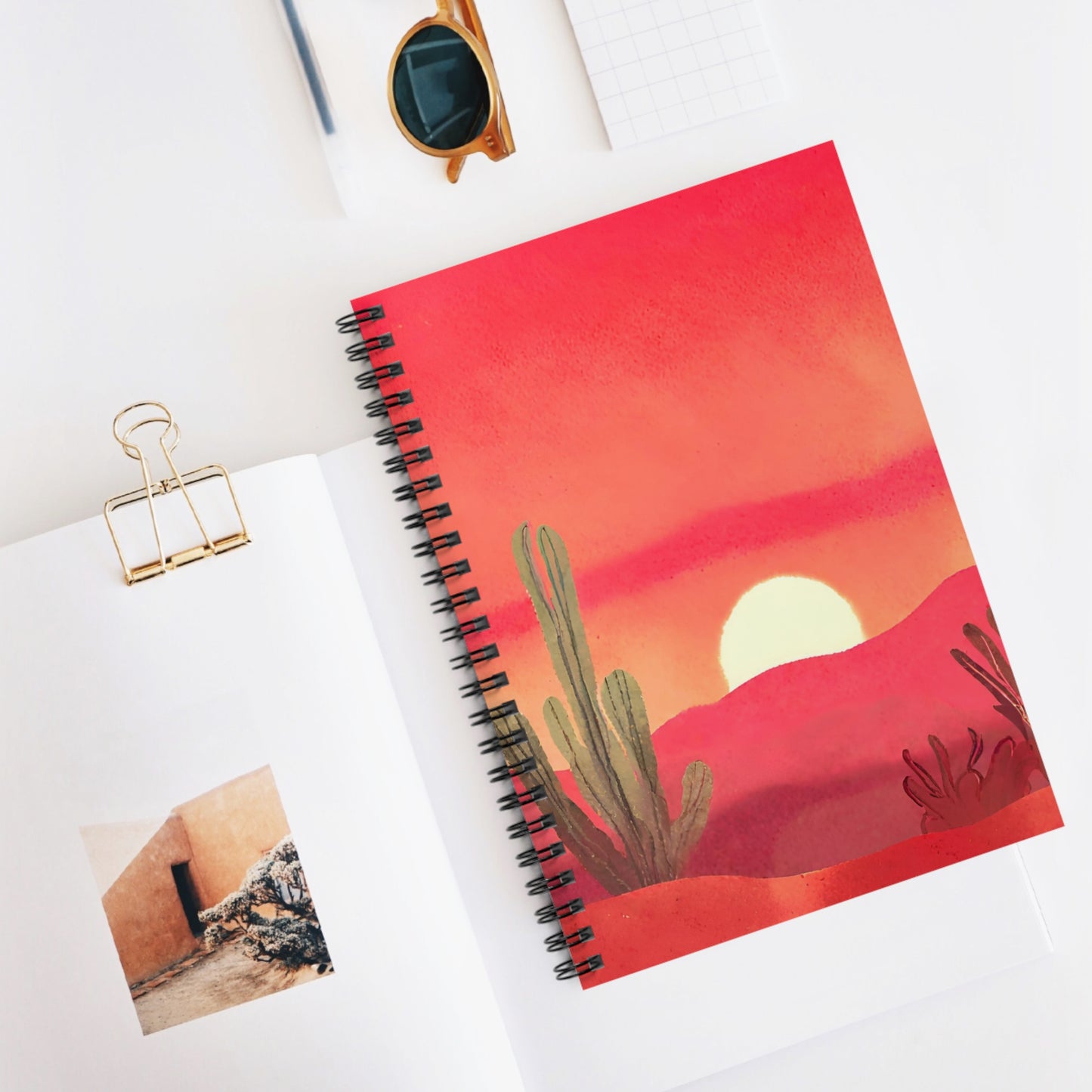 Notebook with desert design on cover, placed on a white surface with sunglasses and a photo clip.