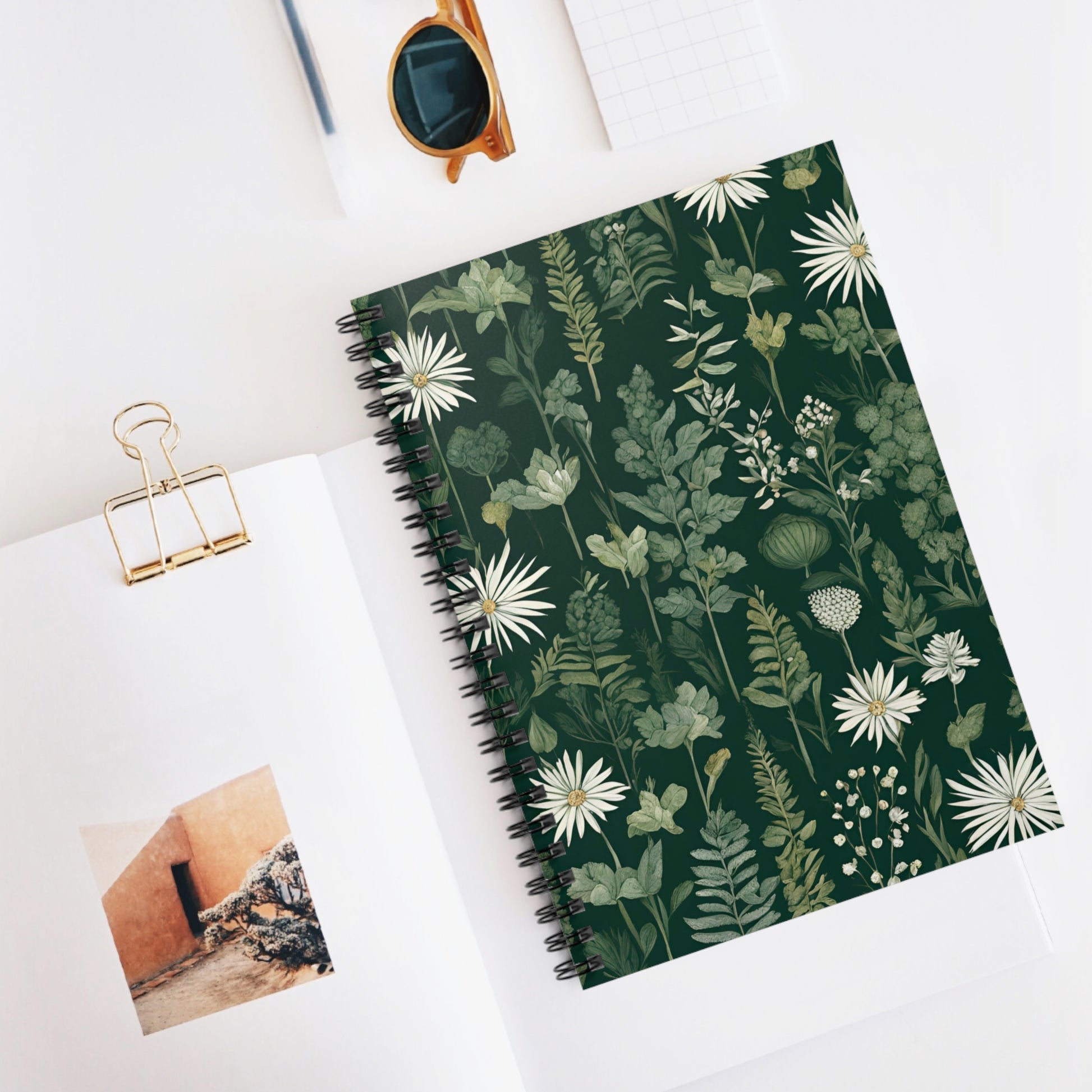 Notebook with floral design on a white surface with sunglasses and a photo clip.