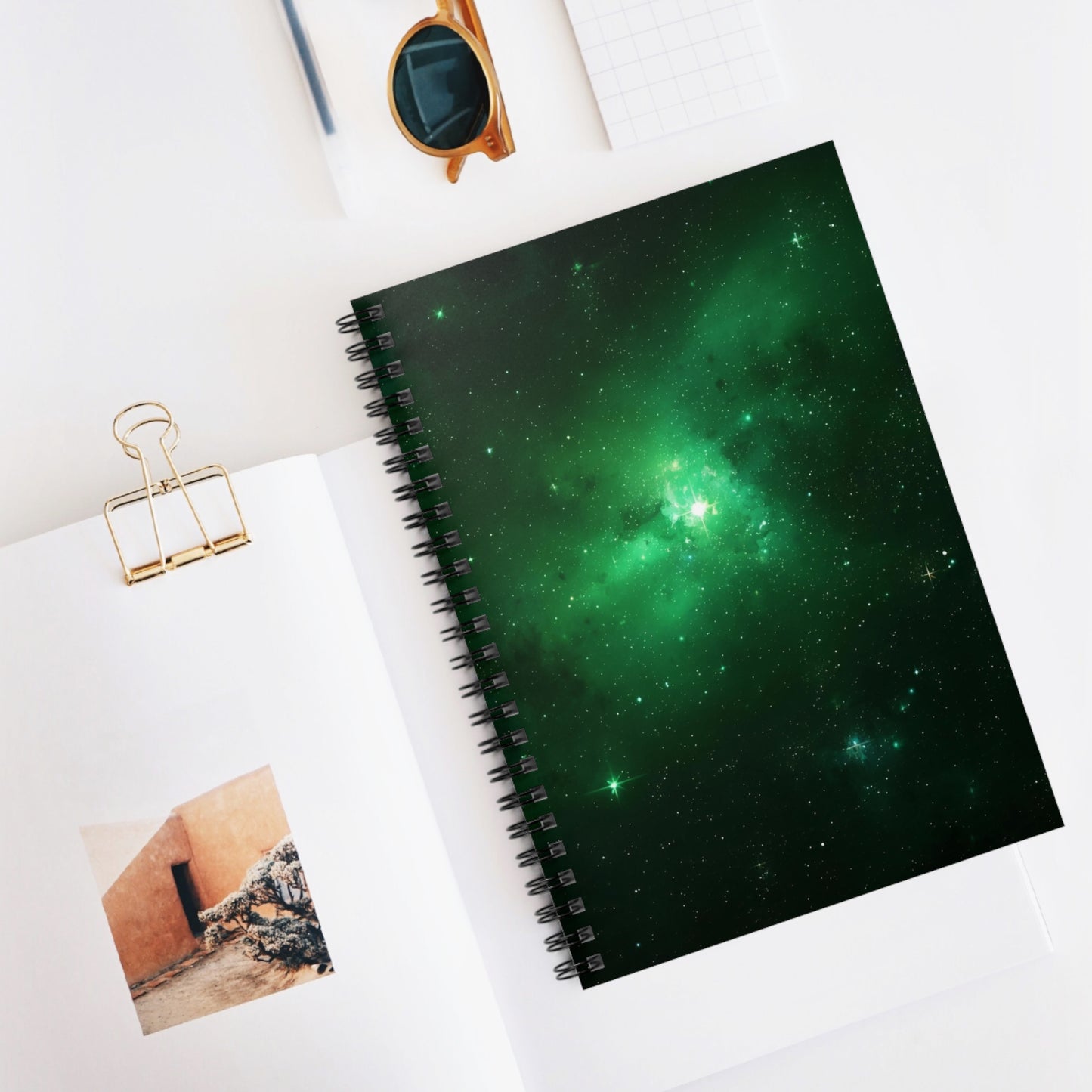 Spiral-bound notebook with green galaxy design on a white surface with sunglasses and a photo clip.