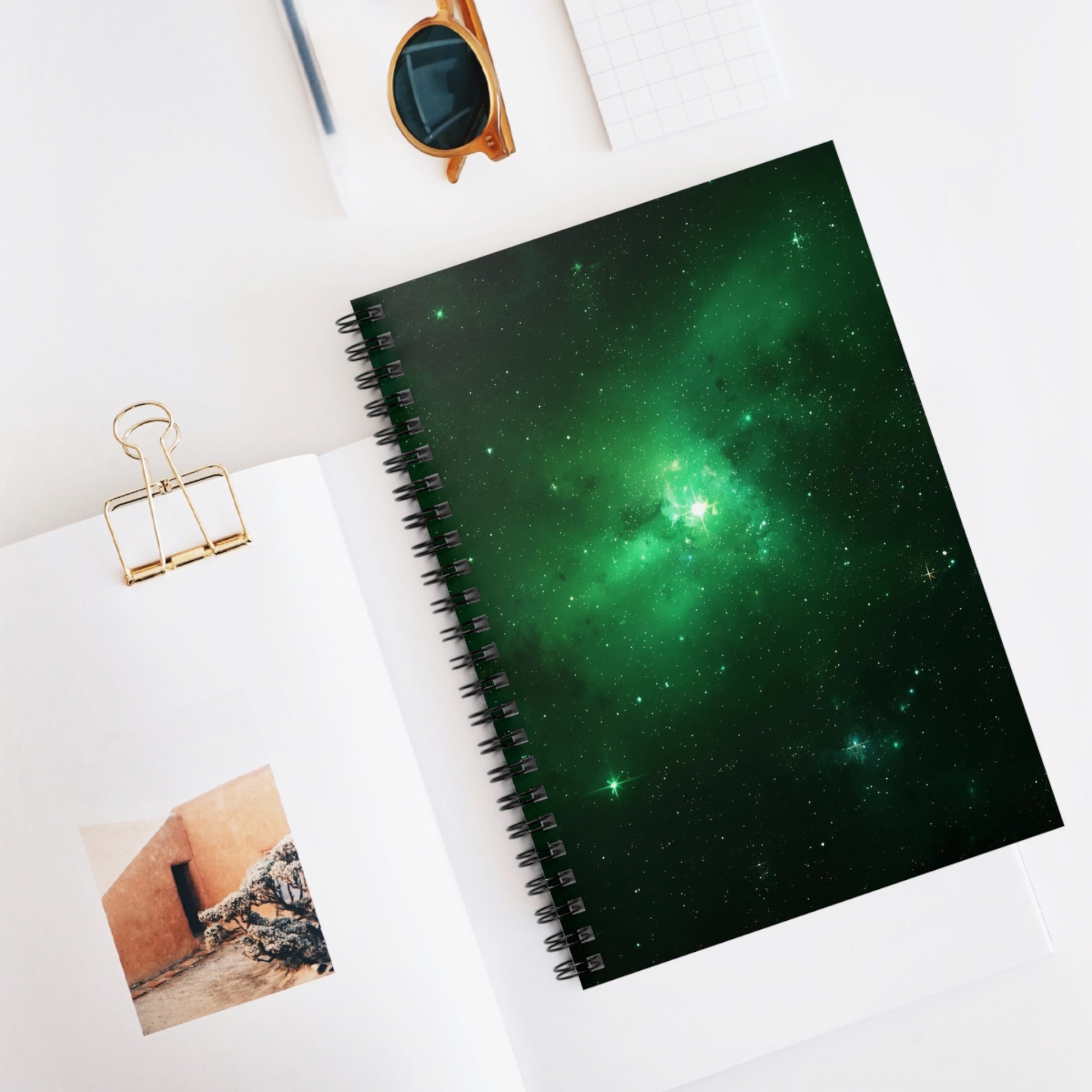 Spiral-bound notebook with green galaxy design on a white surface with sunglasses and a photo clip.