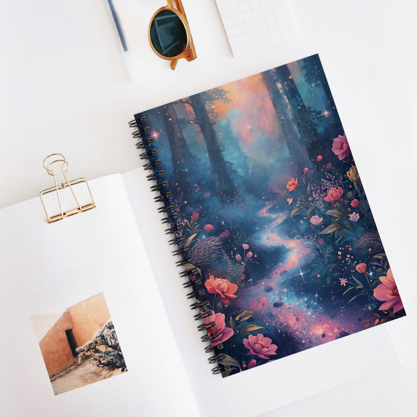 Notebook with a colorful floral and galaxy design on a white surface with a photo clip and sunglasses.