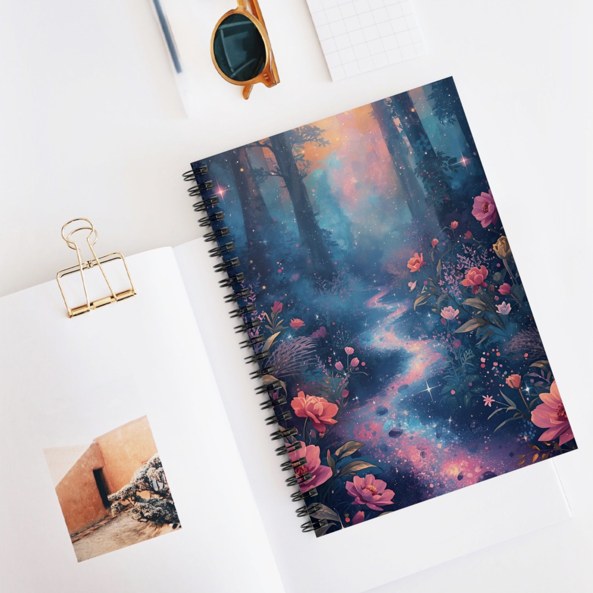 Notebook with a colorful floral and galaxy design on a white surface with a photo clip and sunglasses.