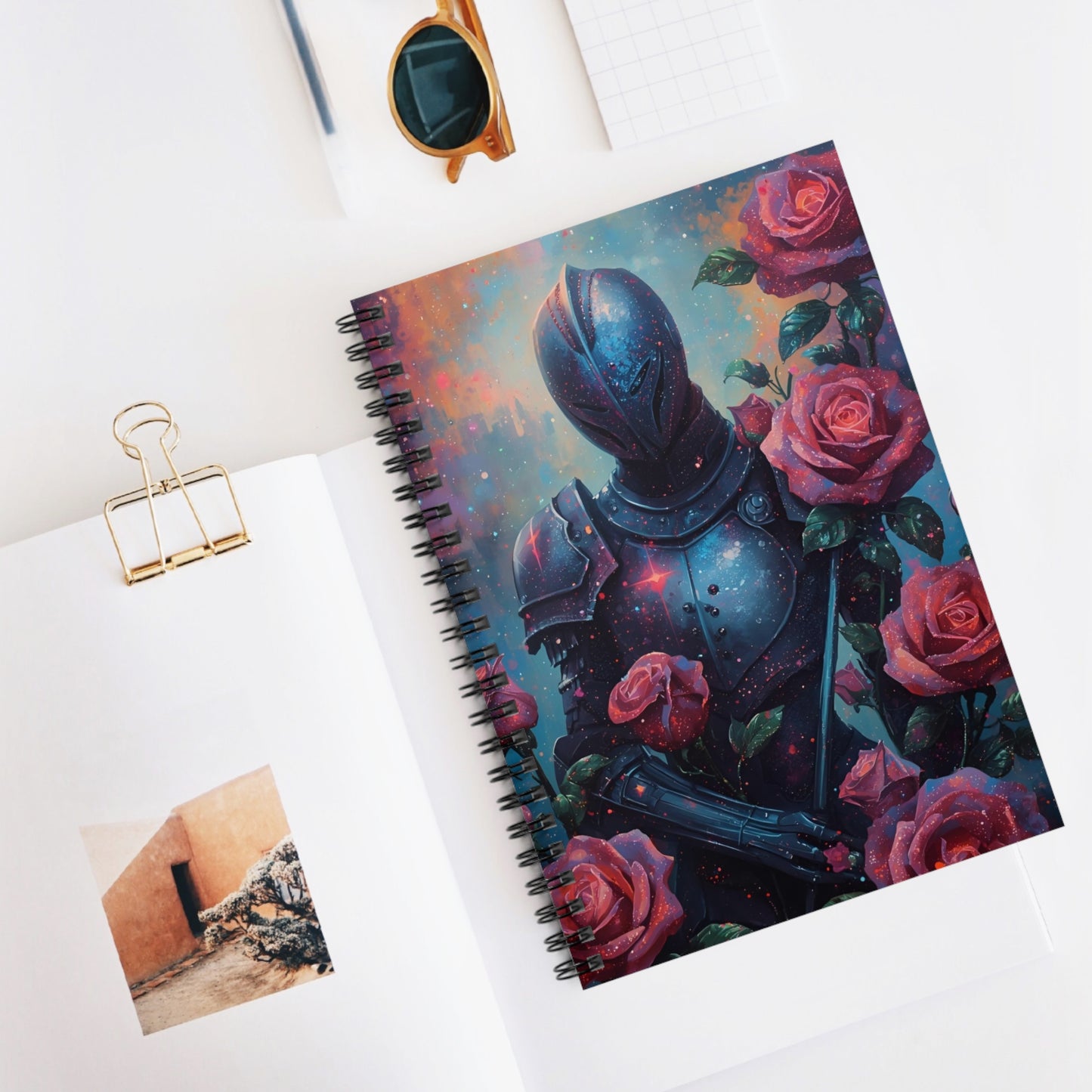 Notebook with a cover design of armor and roses on a white surface with sunglasses and a photo clip.