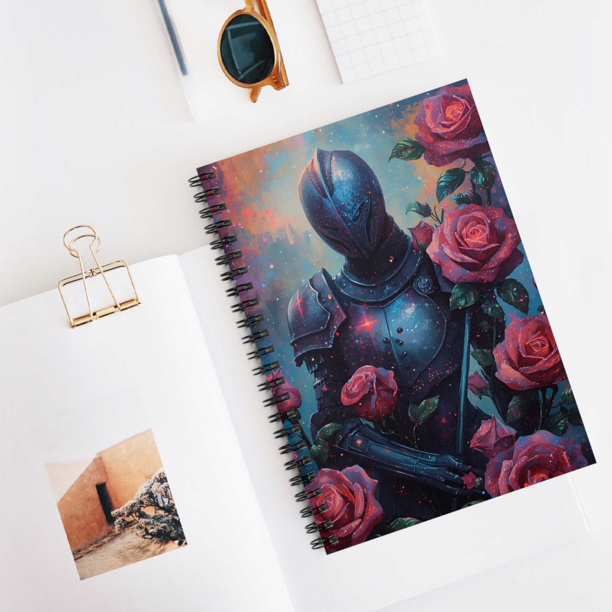 Notebook with a cover design of armor and roses on a white surface with sunglasses and a photo clip.