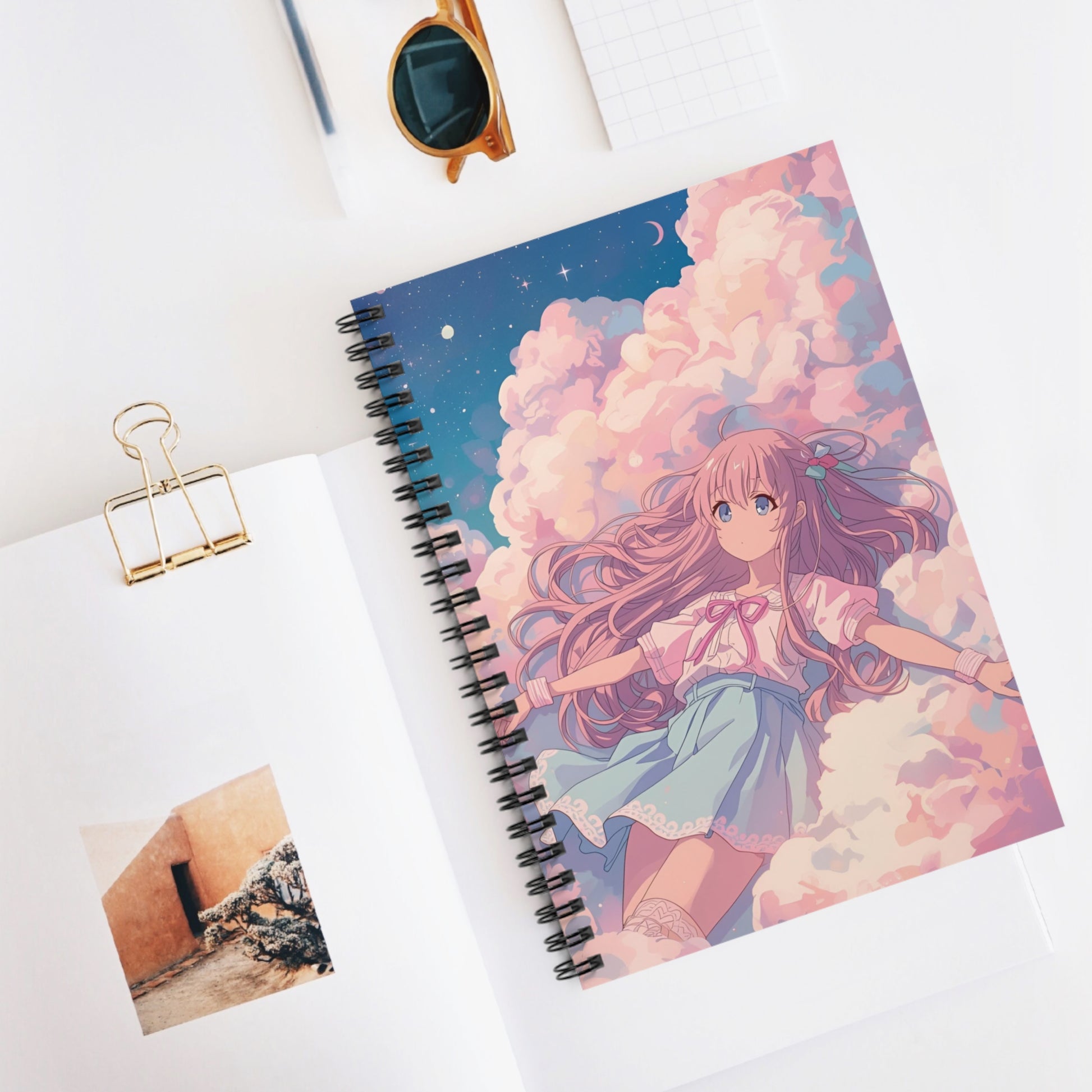 Notebook with an anime-style cover featuring a girl in a cloud, placed on a white surface with sunglasses and a photo clip.