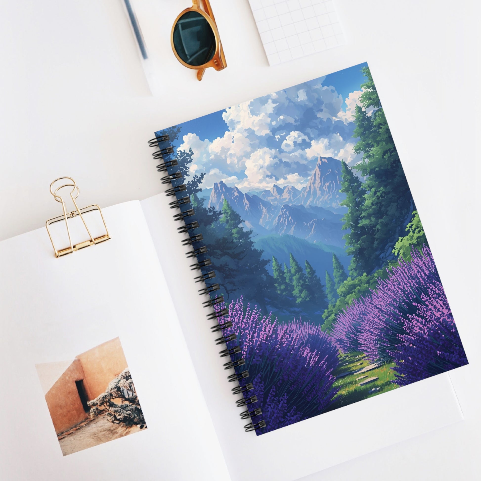 Notebook with a mountain and lavender design on a white surface with sunglasses and a photo clip.