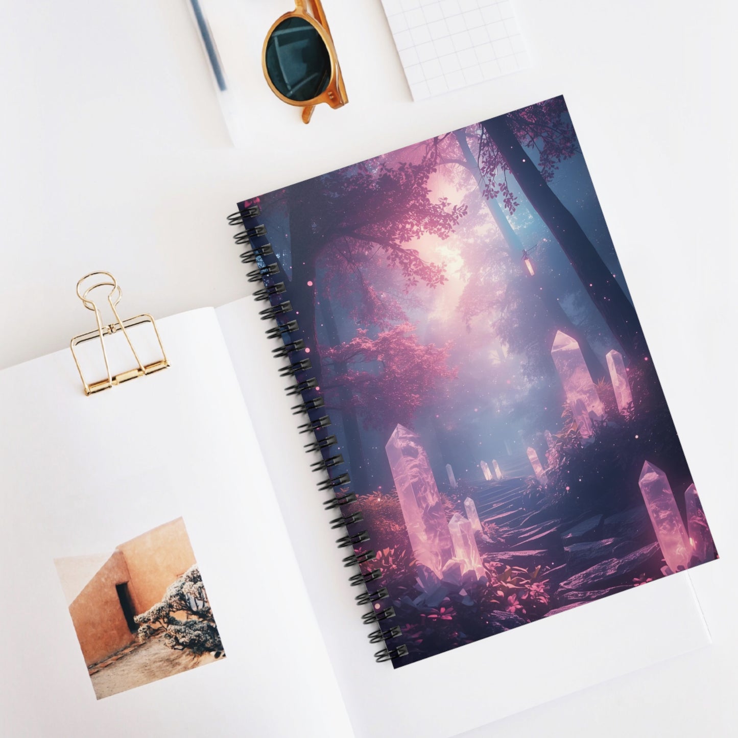 Spiral-bound notebook with a fantasy-themed cover on a white surface, accompanied by sunglasses and a photo clip.