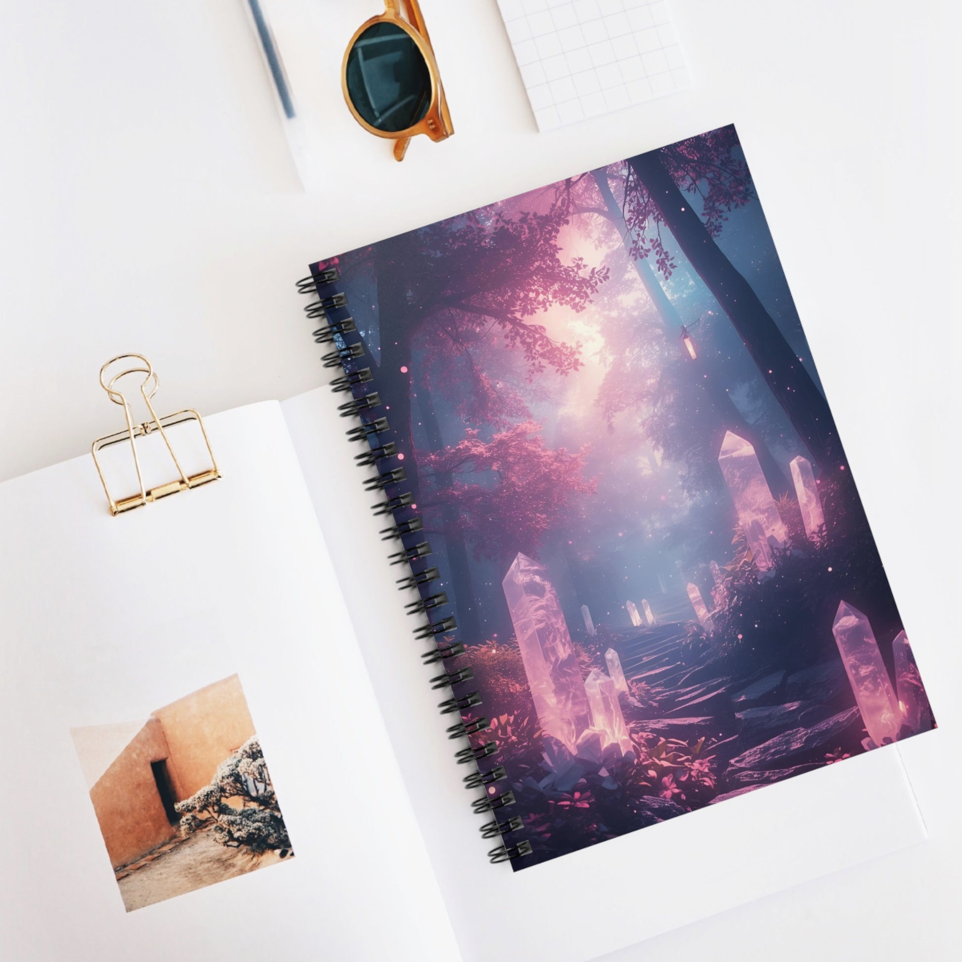Spiral-bound notebook with a fantasy-themed cover on a white surface, accompanied by sunglasses and a photo clip.